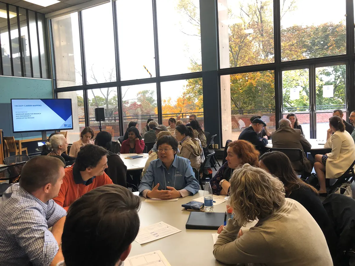 Group of Faculty sitting around tables discussing things with large windows behind them.